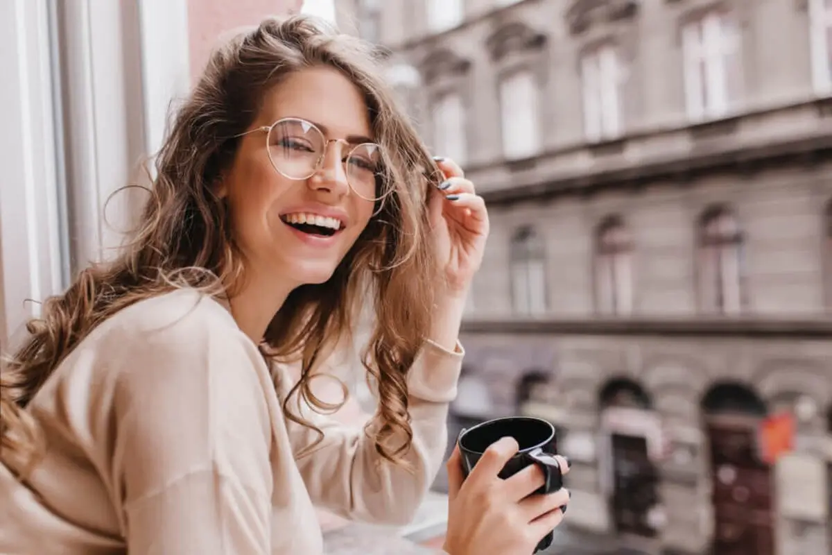 Woman smiling by a window wearing glasses, holding a coffee mug